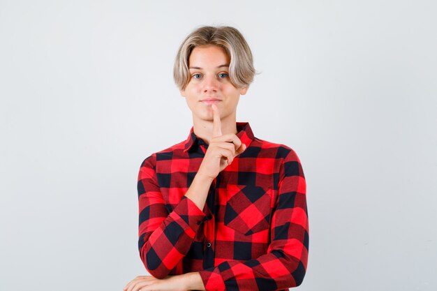 Young teen boy in checked shirt keeping finger on chin and looking confident , front view.