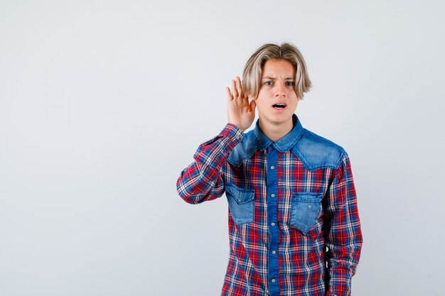 Young teen boy in checked shirt keeping hand behind ear and looking confused , front view.