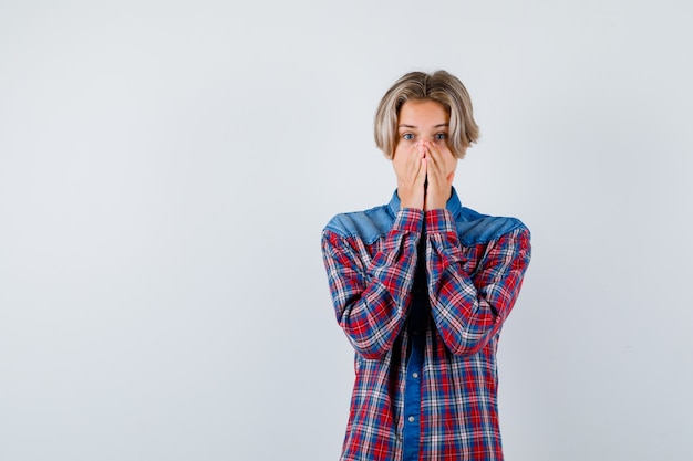 Young teen boy in checked shirt keeping hands on mouth and looking scared , front view.