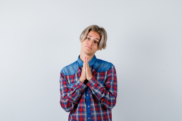チェックシャツを着た若い10代の少年は、祈りのジェスチャーで手を保ち、希望に満ちた正面図を探しています。