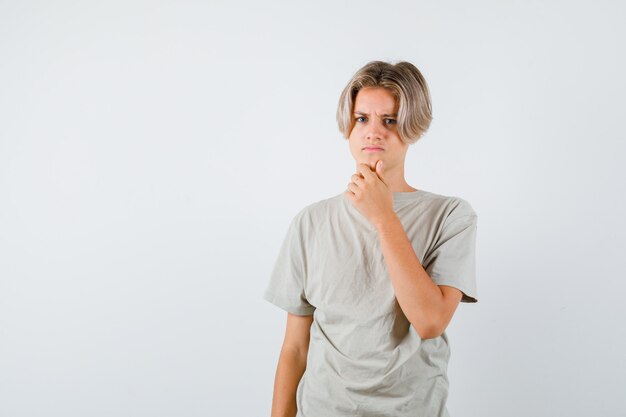 Young teen boy keeping hand on chin in t-shirt and looking upset , front view.