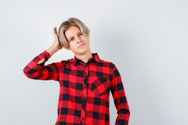 Young teen boy keeping hand on head in checked shirt and looking confident , front view.