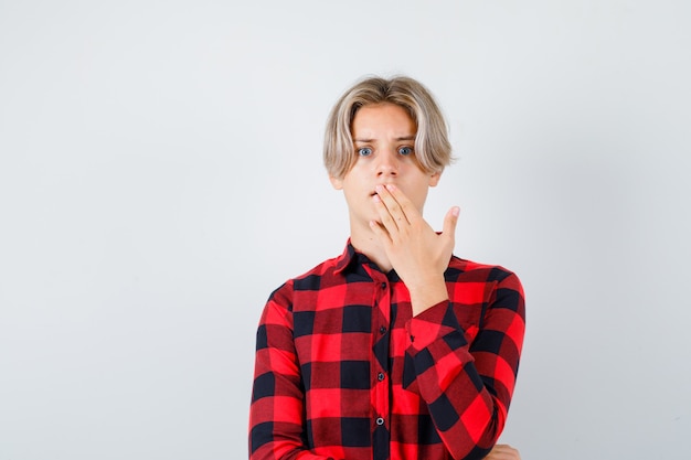 Young teen boy keeping hand on mouth in checked shirt and looking shocked , front view.