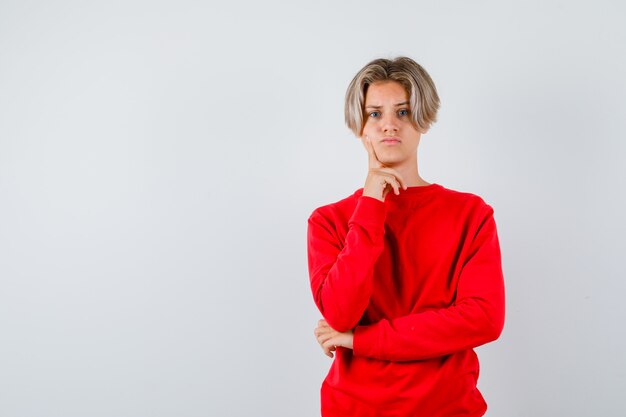 Young teen boy in red sweater holding finger on cheek and looking sad , front view.