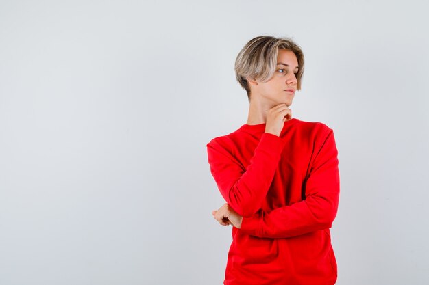 Young teen boy in red sweater looking aside with chin propped on hand and looking focused , front view.