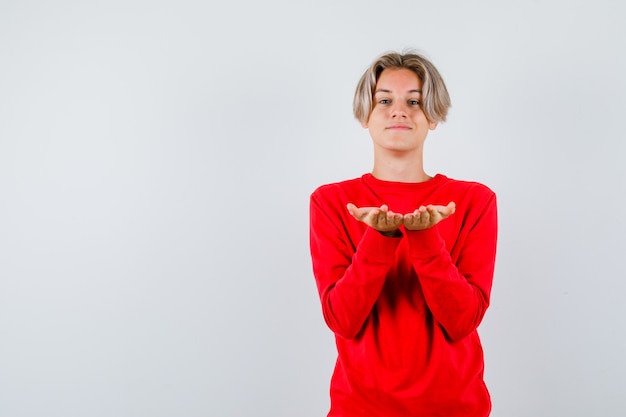 Foto gratuita giovane ragazzo adolescente in maglione rosso che fa il gesto di dare o ricevere e sembra allegro, vista frontale.