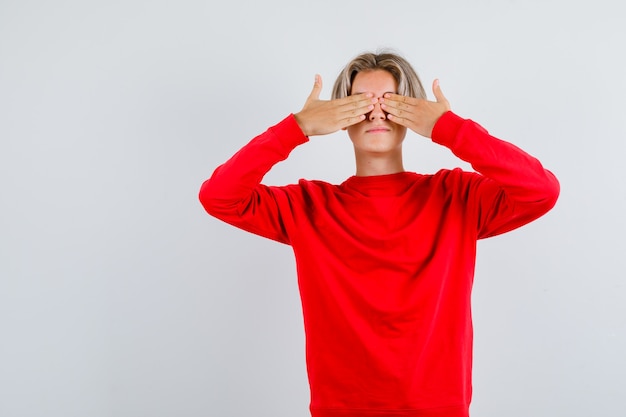 Young teen boy in red sweater with hands on eyes and looking excited , front view.