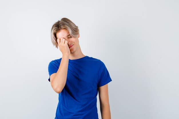 Young teen boy rubbing eye while crying in blue t-shirt and looking depressed. front view.
