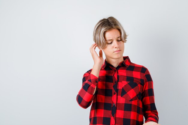 Young teen boy scratching head, looking down in checked shirt and looking pensive , front view.