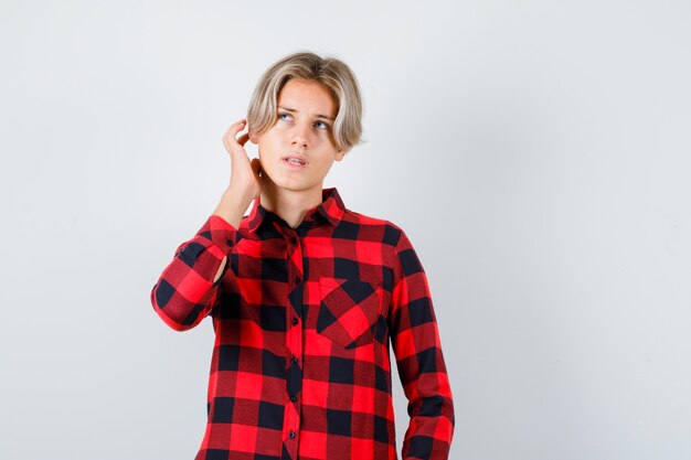 Young teen boy scratching his head in checked shirt and looking preoccupied , front view.