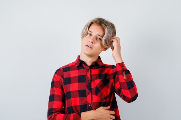 Young teen boy scratching his head in checked shirt and looking thoughtful , front view.