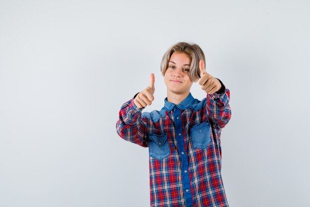 Молодой подросток мальчик показывает двойные пальцы вверх в клетчатой рубашке и выглядит веселым. передний план.