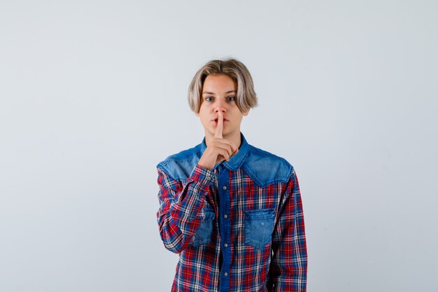 Young teen boy showing silence gesture in checked shirt and looking careful. front view.