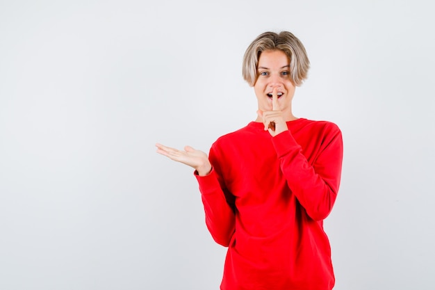 Young teen boy showing silence gesture, spreading palm aside in red sweater and looking cheery , front view.