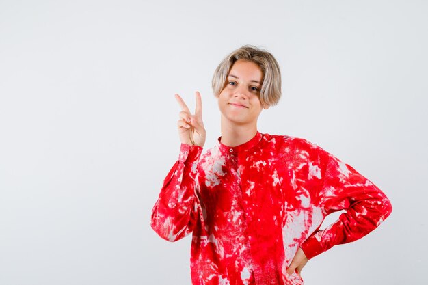 Young teen boy showing victory gesture in shirt and looking cheerful , front view.