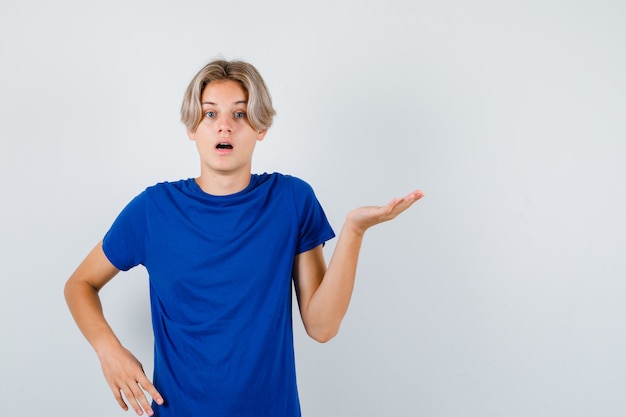 Young teen boy spreading palm aside in blue t-shirt and looking puzzled , front view.