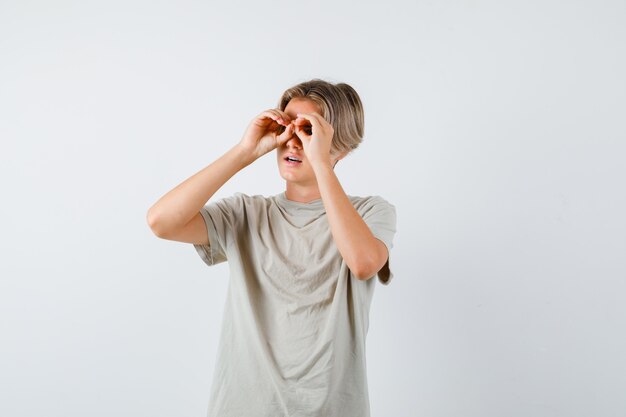 Young teen boy in t-shirt showing glasses gesture while looking away and looking wondered , front view.