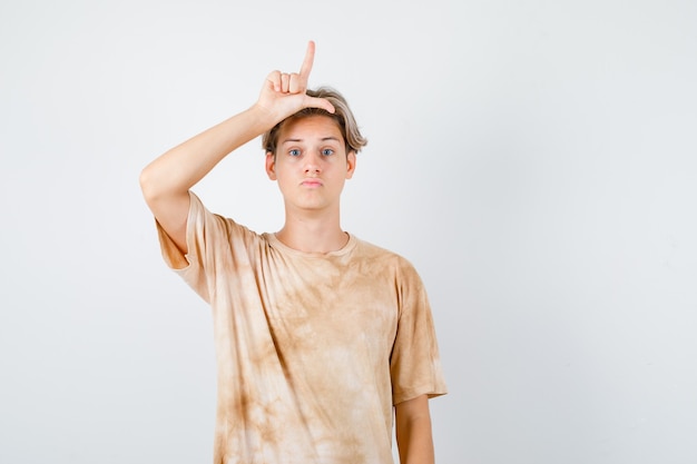 Free photo young teen boy in t-shirt showing loser sign on head and looking disappointed , front view.