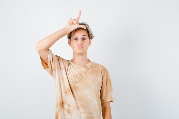Молодой мальчик-подросток в футболке показывает знак проигравшего на голове и выглядит разочарованным, вид спереди.