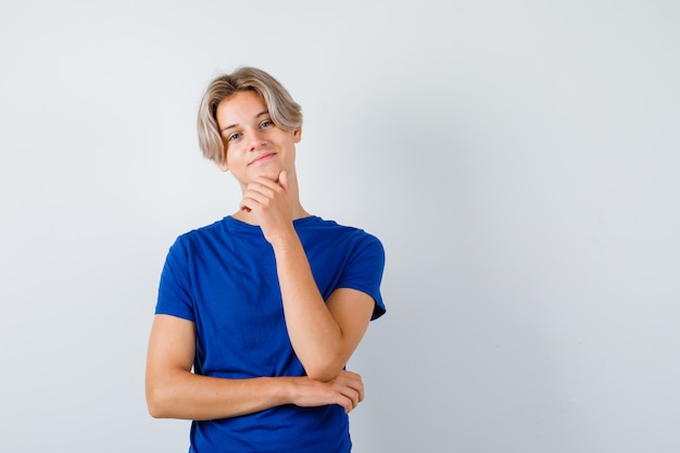 Young teen boy with hand on chin in blue t-shirt and looking jovial , front view.