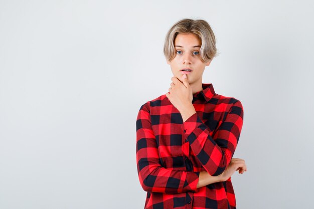 Young teen boy with hand on chin in checked shirt and looking puzzled , front view.