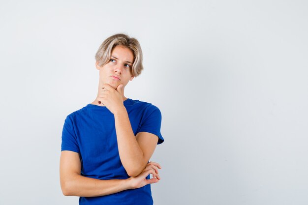 Young teen boy with hand on chin, looking up in blue t-shirt and looking thoughtful. front view.