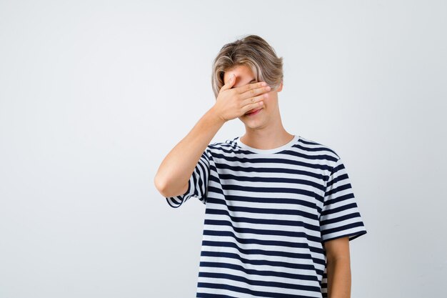 縞模様のTシャツを着て、恥ずかしそうに見える顔に手を持っている若い10代の少年。正面図。