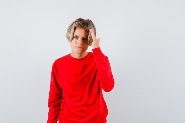 Young teen boy with hand over head to see clearly in red sweater and looking confused , front view.