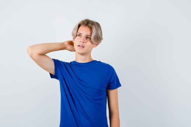 Young teen boy with hand behind head while looking away in blue t-shirt and looking pensive , front view.