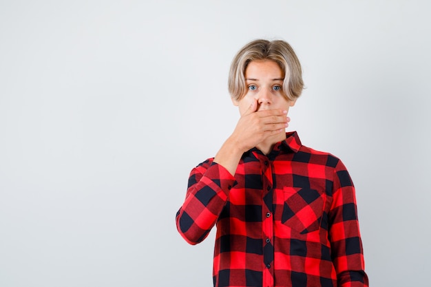 Free photo young teen boy with hand on mouth in checked shirt and looking surprised , front view.