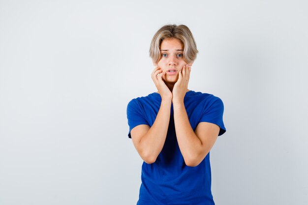 Young teen boy with hands on cheeks in blue t-shirt and looking anxious , front view.