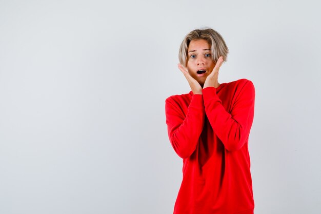 Young teen boy with hands on cheeks in red sweater and looking frightened , front view.