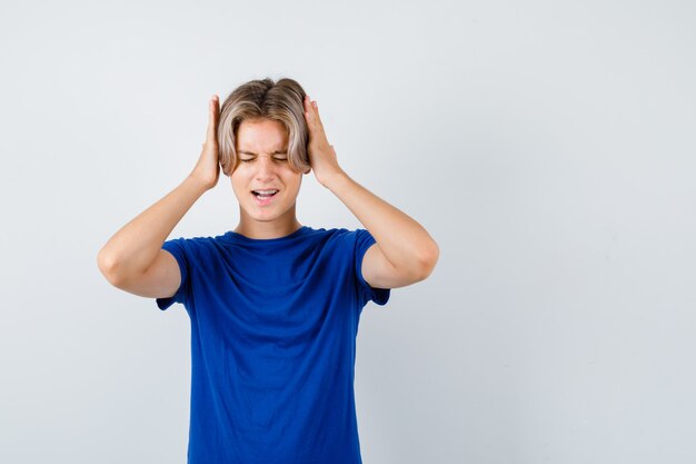 Young teen boy with hands on ears in blue t-shirt and looking annoyed , front view.