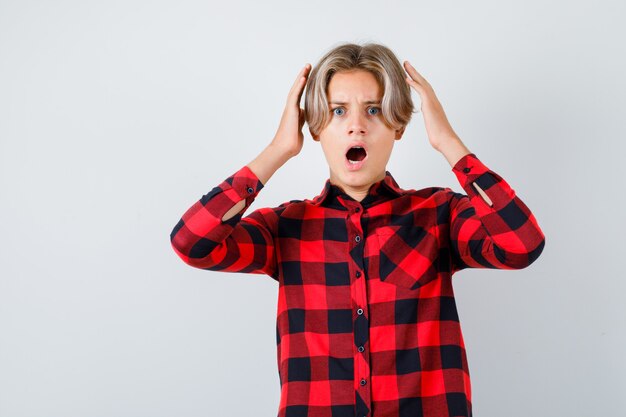 Young teen boy with hands near head in checked shirt and looking agitated , front view.