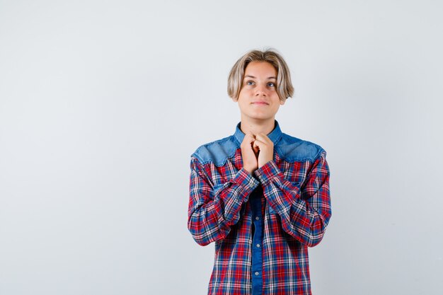 Молодой подросток мальчик с руками в молитвенном жесте в клетчатой рубашке и выглядит обнадеживающим