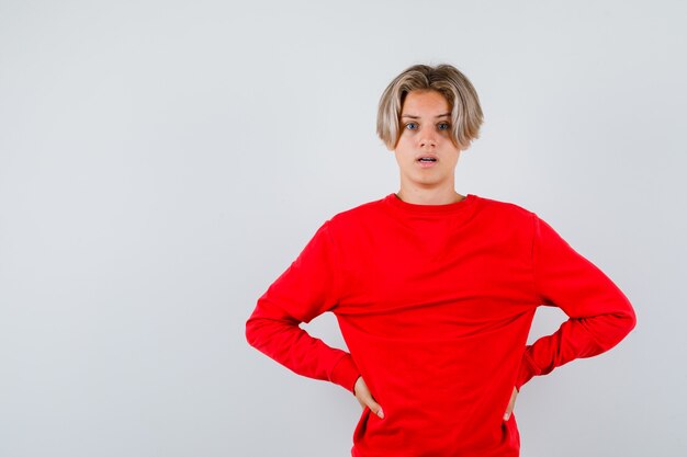 Young teen boy with hands on waist in red sweater and looking bewildered , front view.