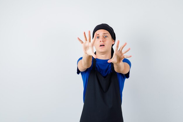 Young teen cook in t-shirt, apron showing stop gesture and looking terrified , front view.