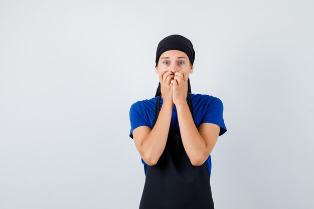 Young teen cook in t-shirt, apron with hands on mouth and looking terrified , front view.