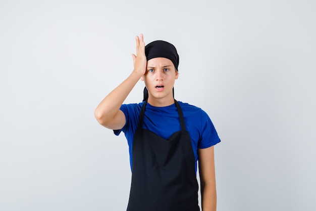 Молодой подросток готовит с рукой на голове в футболке, фартуке и смотрит задумчиво, вид спереди.