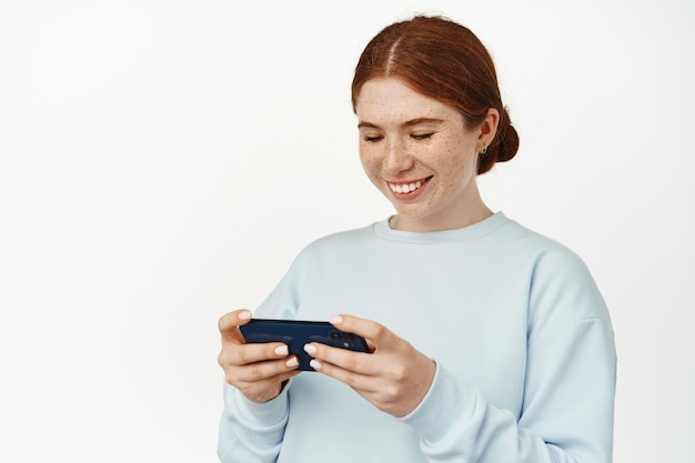 Giovane ragazza dai capelli rossi che gioca ai videogiochi sul suo telefono su bianco