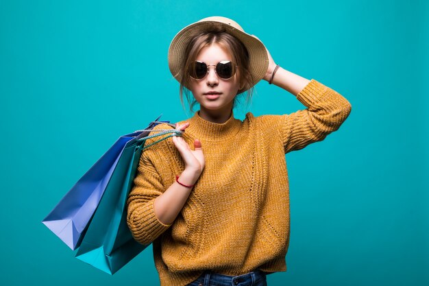 Young teen woman in sunglasses and hat holding shopping bags in her hands feeling so happiness isolated on green wall