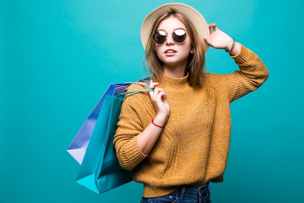 Young teen woman in sunglasses and hat holding shopping bags in her hands feeling so happiness isolated on green wall