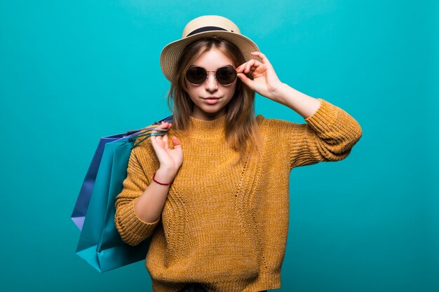 Young teen woman in sunglasses and hat holding shopping bags in her hands feeling so happiness isolated on green wall
