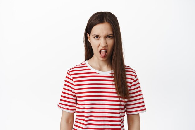 Young teenage girl show dislike, extend tongue, frowning and squinting from disgust, aversion, bad taste of smth awful, standing in t-shirt against white background.