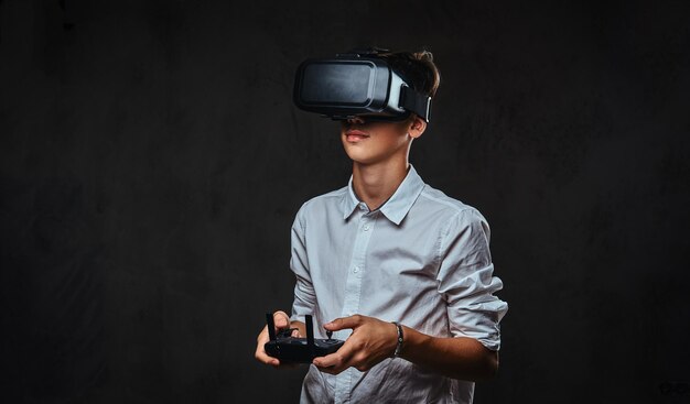 Молодой подросток, одетый в белую рубашку, носит очки виртуальной реальности и управляет квадрокоптером с помощью пульта дистанционного управления.