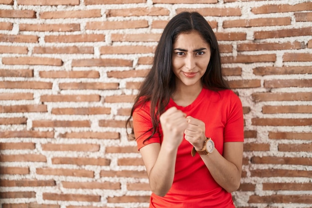 Young teenager girl standing over bricks wall ready to fight with fist defense gesture angry and upset face afraid of problem