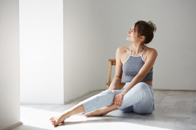 Young tender beautiful woman smiling looking at window sitting on floor over white wall.