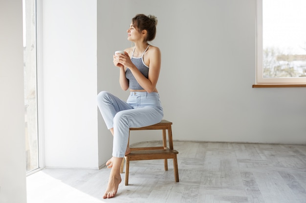 Free photo young tender brunette woman smiling holding cup looking at window enjoying view sitting on chair over white wall in morning.