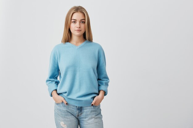 Young tender fair haired teenage girl with healthy skin wearing blue top looking  with serious or pensive expression. Caucasian woman model with hands in pockets posing indoors
