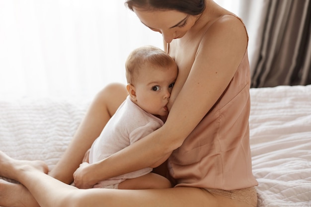아침에 침대에 앉아 그녀의 신생아를 포옹 잠옷 모유 수유에 젊은 부드러운 어머니